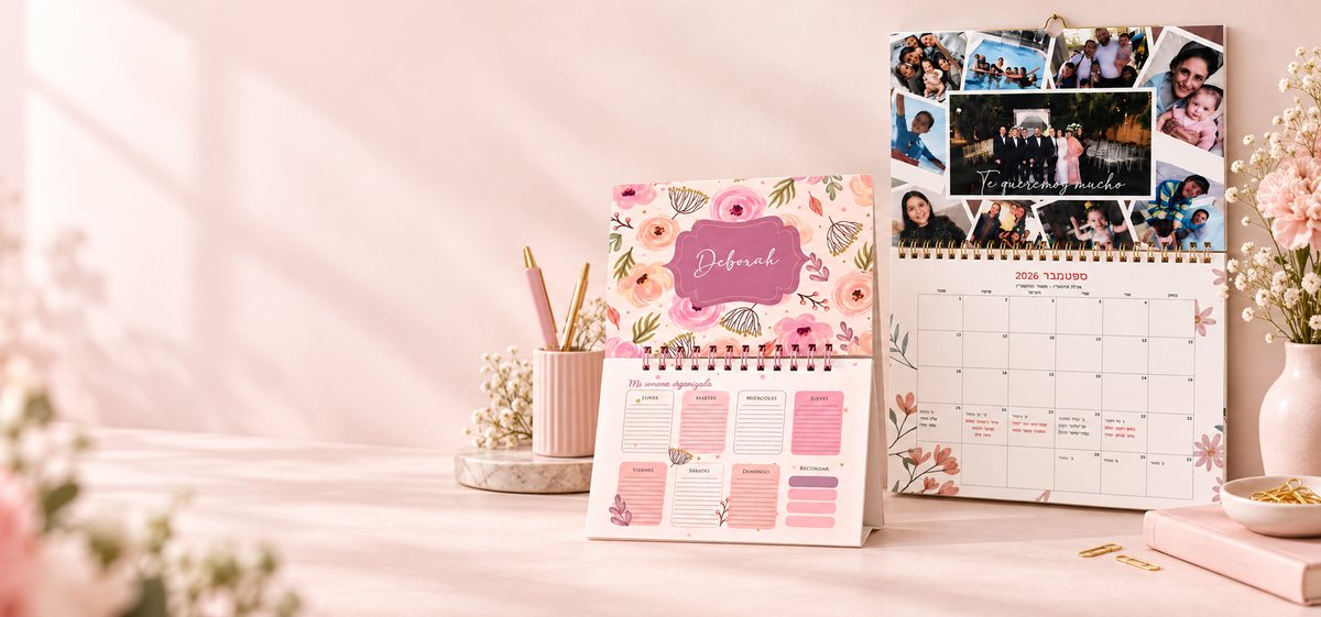 A pink floral desk calendar with a photo collage page and planner inserts displayed on a light pink surface. The scene includes a matching pink cup with pens, small flowers, and a vase in a softly styled interior setup.